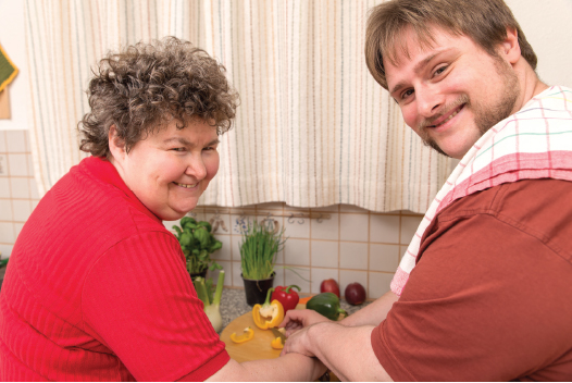 Man and woman smiling at camera as they cut up fresh vegetables.