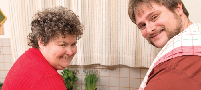 Man and woman smiling at camera as they cut up fresh vegetables.