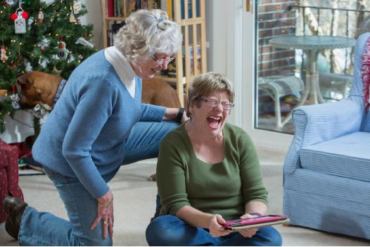 Women laughing at Christmas as one of them plays with tablet and the other watches. 